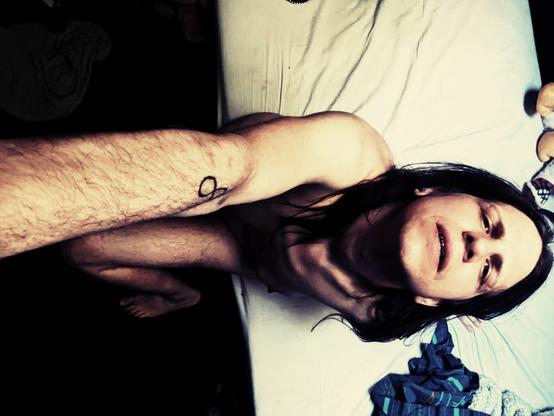 Void sits nude on the edge of bed, face tilted back to stare up at the camera it holds above it. It has long hair, prominent collar bones and hairy, lanky limbs.